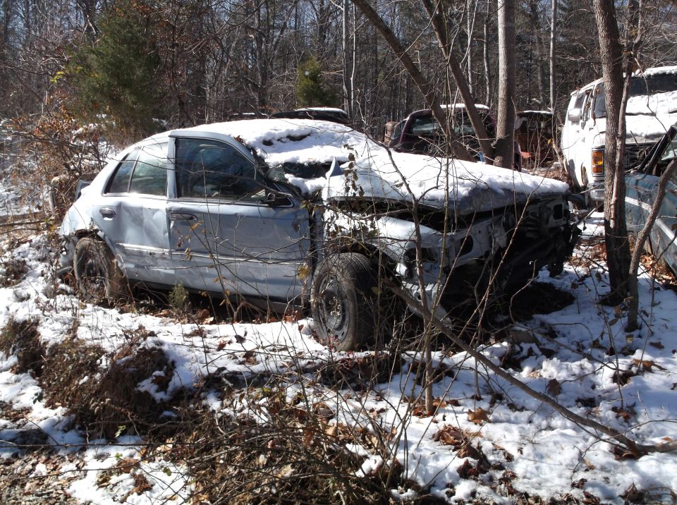 Damaged light blue sedan in a snowy, wooded area with front end missing.