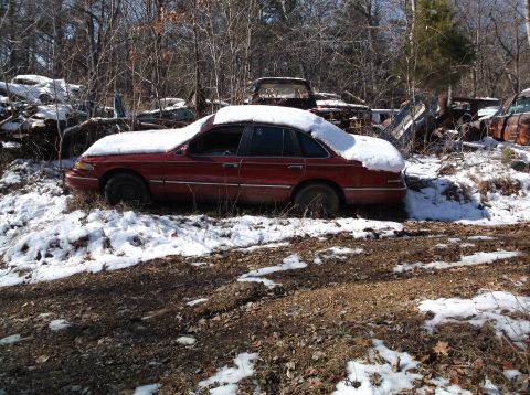 1974 Dodge Challenger - A red sedan covered in snow sits in a junkyard surrounded by trees and other discarded vehicles. The car appears to be a late 1990s model w…