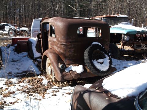1974 Dodge Challenger - A rusty, vintage sedan sits in a snowy junkyard, with a partial view of a turquoise and white truck behind it.