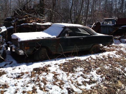 1974 Dodge Challenger - A dark green, four-door sedan sits in a snowy junkyard with a pile of scrap metal behind it and a blue truck in the background.