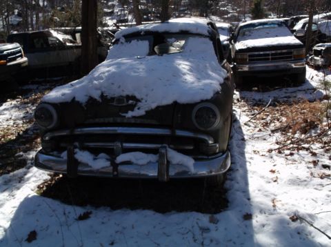 1974 Dodge Challenger - A front view of a dark-colored, older model car covered in snow, parked outdoors in a wooded area. The vehicle has a chrome grille and bump…