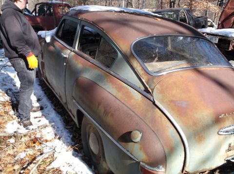 1974 Dodge Challenger - The rear passenger side of a heavily rusted, vintage four-door sedan sits in a snowy, wooded area. A person in a black hoodie and yellow gl…