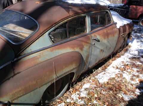 1974 Dodge Challenger - The rear passenger side of a heavily rusted, vintage sedan is shown, covered in patches of snow and surrounded by fallen leaves. The car's…
