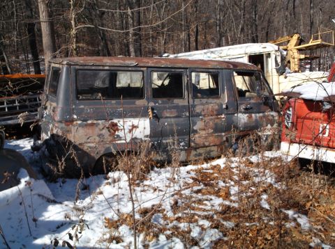 1974 Dodge Challenger - A heavily rusted, dark-colored van sits in a snowy, wooded junkyard next to other vehicles and equipment. The van has multiple windows and…