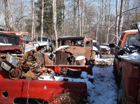 1974 Dodge Challenger - A collection of old, rusted vehicles are parked in a snowy wooded area. The foreground shows parts of several trucks, including a red truck…