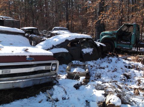 1974 Dodge Challenger - A collection of old vehicles, covered in snow, sit in a wooded area. A red pickup truck is in the foreground, with a dark-colored sedan and…