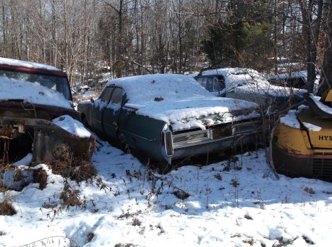 1974 Dodge Challenger - A green sedan covered in snow sits in a junkyard with other vehicles and trees in the background. The rear of the car is visible, showing i…