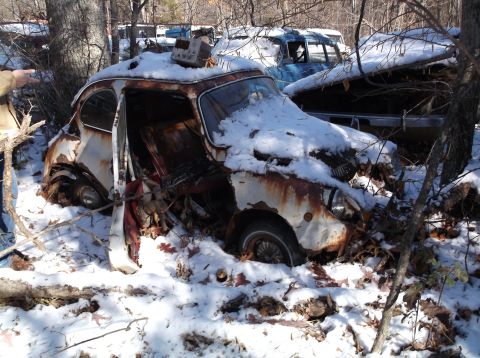 1974 Dodge Challenger - A heavily rusted and snow-covered vintage car sits in a wooded junkyard. The driver's side door is open, revealing the interior, and a smal…
