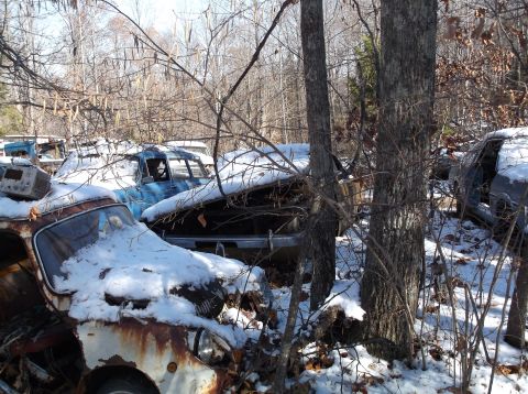 1974 Dodge Challenger - Several old cars are parked in a wooded area, covered in snow. The foreground shows the front of a rusty, white vehicle, while a blue sedan…