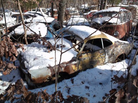 1974 Dodge Challenger - A rusty, snow-covered vintage car is partially visible in a wooded junkyard setting. Another older vehicle, possibly a van, is visible behi…