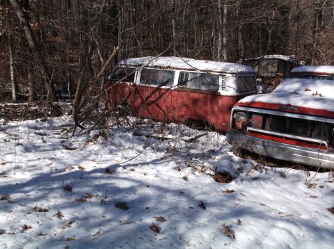 1974 Dodge Challenger - Two abandoned vehicles sit in a snowy wooded area. A red and white van is partially visible on the left, and the front of a red Dodge picku…