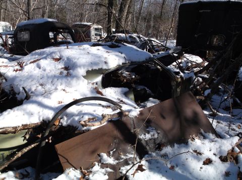1974 Dodge Challenger - A pile of rusted car parts and vehicle shells are covered in snow and fallen leaves in a wooded area. The scene includes a dark-colored car…