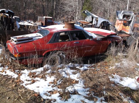 1974 Dodge Challenger - A red coupe sits in a junkyard with snow on the ground and a rusty barrel on its roof. Other wrecked vehicles are visible in the background.