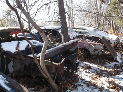 1974 Dodge Challenger - Several old, rusted-out cars are partially covered in snow and debris in a wooded area. The vehicles appear to be from the 1970s, with one…