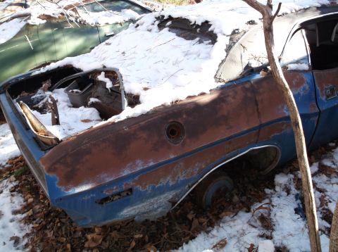1974 Dodge Challenger - A heavily rusted blue 1974 Dodge Challenger sits abandoned in the snow, with its trunk open and interior exposed to the elements. Another v…