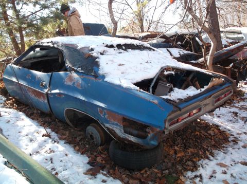 1974 Dodge Challenger - A weathered blue 1974 Dodge Challenger coupe sits in a snowy junkyard, its roof and trunk covered in snow. The car is missing its wheels an…
