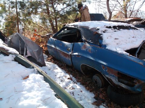 1974 Dodge Challenger - A blue classic car, likely a Dodge Challenger, sits partially covered in snow and fallen leaves in an outdoor junkyard setting. A person in…