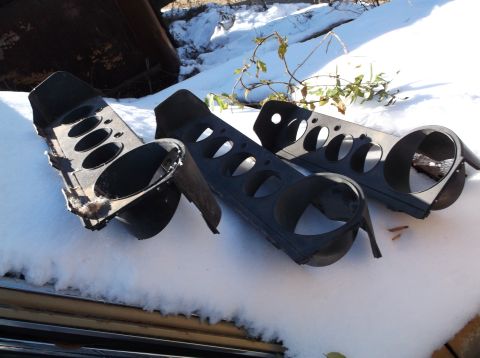 1974 Dodge Challenger - Three black plastic headlight bezels or similar automotive trim pieces are scattered on a snowy surface. The pieces have multiple circular…