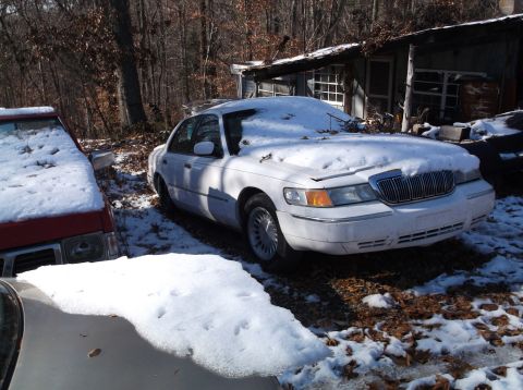 1974 Dodge Challenger - A white sedan covered in snow sits in a wooded area next to a dilapidated structure. Another vehicle, partially covered in snow, is parked…