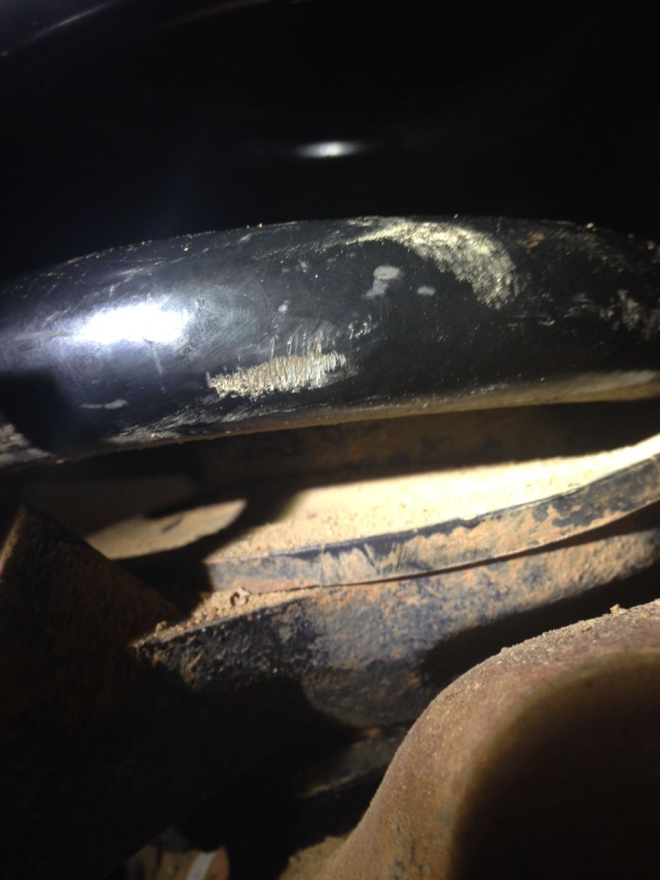 Scuffed black metal suspension part on a vehicle undercarriage, covered in dust.
