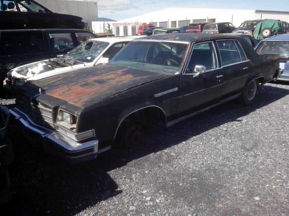 Black four-door sedan with rust on hood, missing front wheel, in a gravel lot.