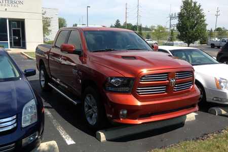 1982 Ford Bronco - A bright orange Dodge Ram pickup truck is parked in a parking lot next to a dark blue sedan and a white sedan. The truck is facing forward…