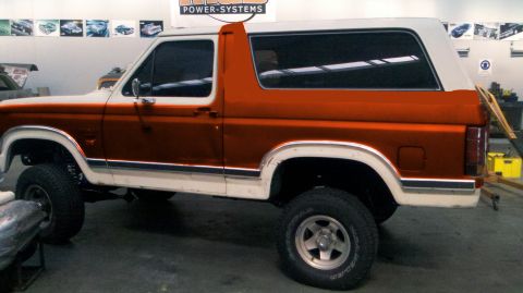 1982 Ford Bronco - The side view of a white and orange two-tone Ford Bronco is shown in a garage setting. The vehicle has chrome trim along the body and fende…