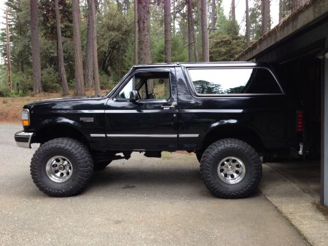 1993 Ford Bronco - A black 1993 Ford Bronco is parked on a paved surface next to a garage. The vehicle has large off-road tires and chrome rims.