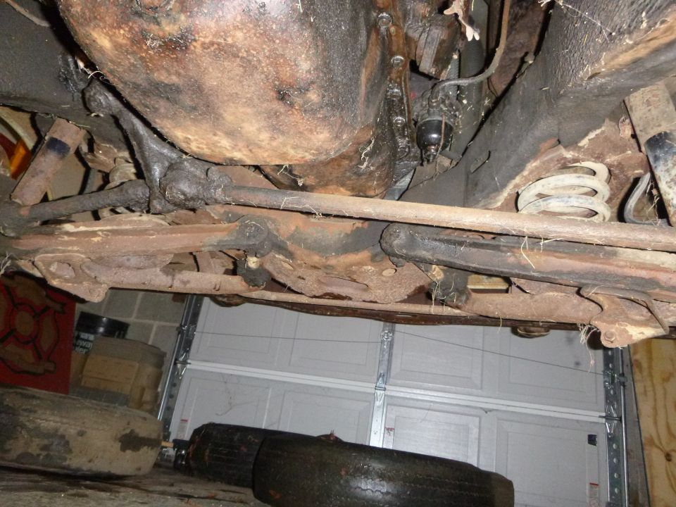 Underside view of a car's suspension and drivetrain components, showing rust and grime. Garage door in background