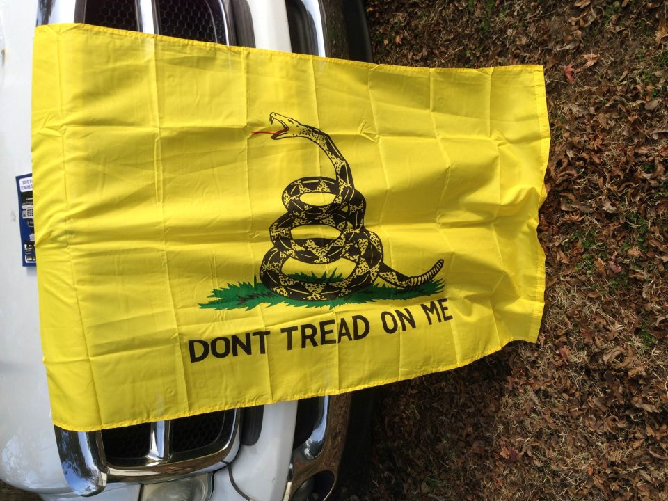 Yellow Gadsden flag with coiled snake and 'DONT TREAD ON ME' text attached to the rear of a white vehicle