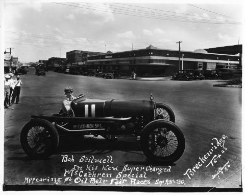 1909 Ford Model T - A driver sits in a vintage race car on a street in front of buildings, with text indicating it's a McCathren Special.