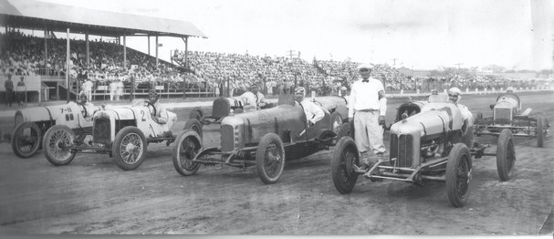 Several old race cars lined up on a track with a grandstand in the background