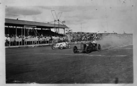 1909 Ford Model T - Race cars speed around a track with a packed grandstand watching the event.