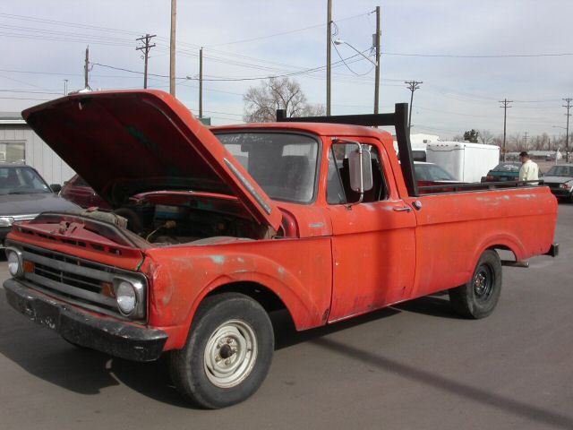 Red 1961 Ford F-100 pickup with hood open and worn paint, parked in a lot