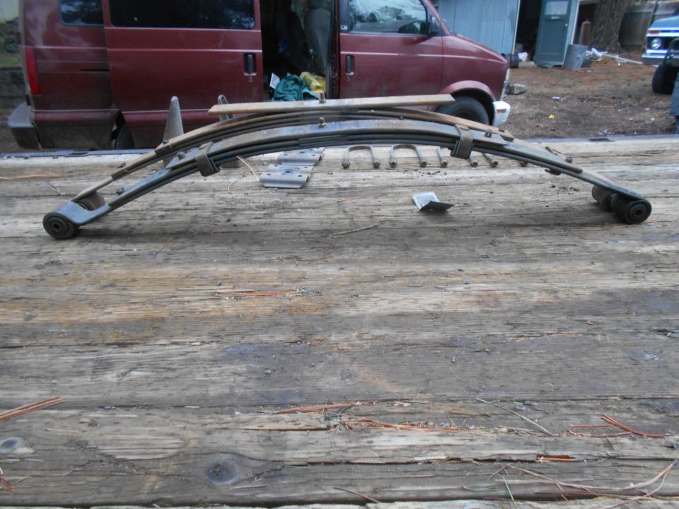 Leaf spring assembly with U-bolts on wooden surface, maroon van in background