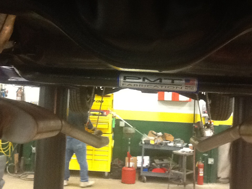 Underside of car on lift showing dual exhaust and a PMT Fabrication badge on a crossmember