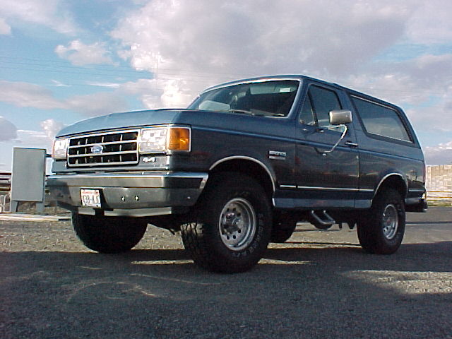 Dark blue Ford Bronco SUV, front three-quarter view, chrome grille and bumper, on gravel under cloudy sky