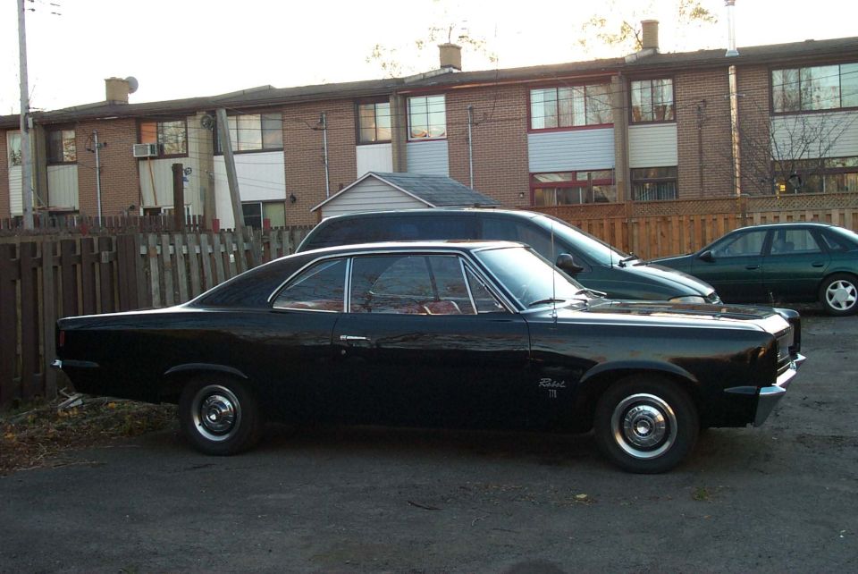 Black coupe parked next to a dark green sedan and a dark SUV. Apartment buildings in background