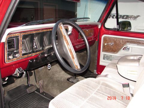 1979 Ford F-150 - The interior of a red 1979 Ford F-150 pickup truck is shown, focusing on the dashboard, steering wheel, and driver's side door panel. Wood…