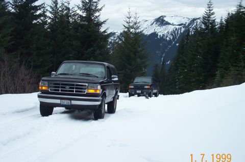 1995 Ford Bronco - Two SUVs navigate a snowy mountain pass, surrounded by dense pine forests and snow-capped peaks.