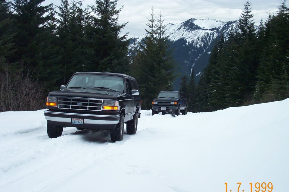 Black Ford Bronco and pickup on a snow-covered mountain road with peaks behind, 1999 photo