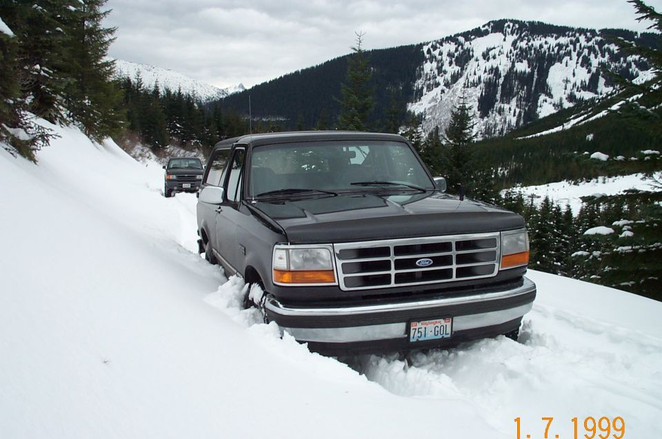 Gray and black Ford Bronco buried in deep snow on a mountain road, front three-quarter view, 1999 date stamp
