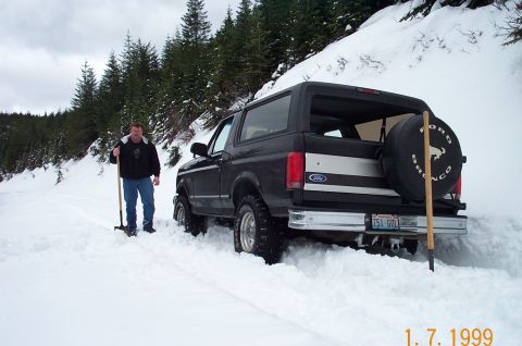 1995 Ford Bronco - A man stands by a black 1995 Ford Bronco partially buried in snow on a wooded hillside.