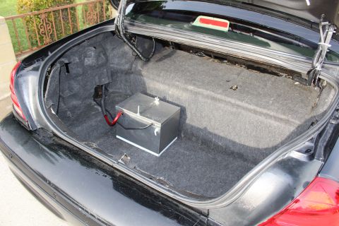 2003 Ford Crown Victoria - open sedan trunk lined with gray carpet, holding a metal battery box with a red positive cable.
