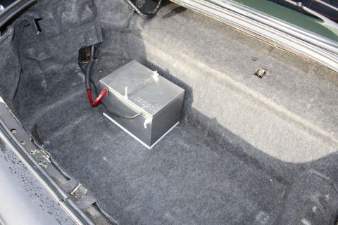 2003 Ford Crown Victoria - Close-up of a metal battery box secured in a carpeted trunk, fed by a red positive cable.