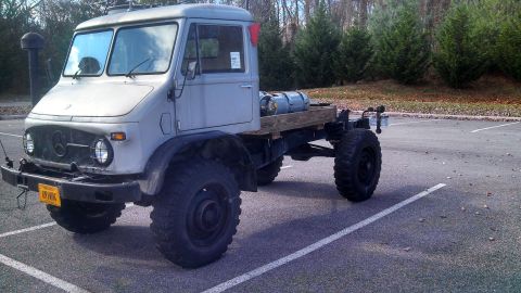 1979 Ford Bronco - A light gray Mercedes-Benz Unimog truck with large off-road tires is parked in a paved lot with trees in the background. The truck has a wo…