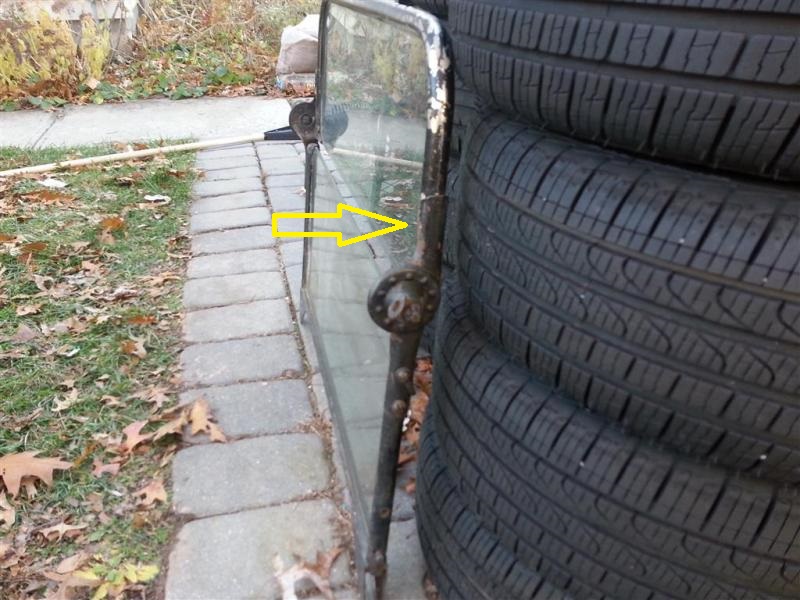 Old Model T windshield frame with glass leaning against stacked tires; yellow arrow points to rusty frame area