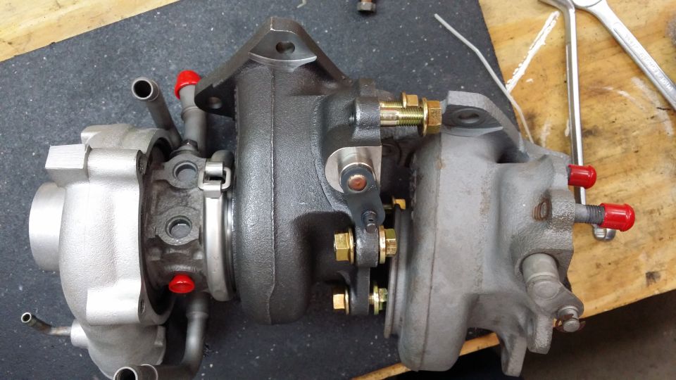 Twin-turbocharger assembly with silver and dark gray housings, brass bolts, and red caps.