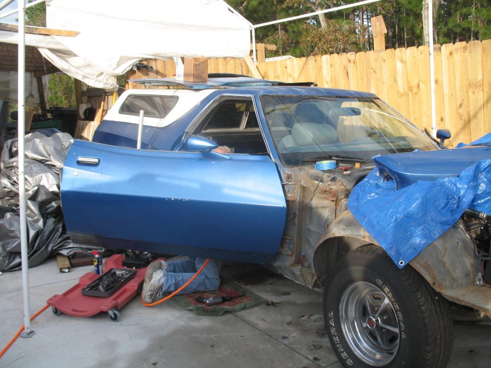 Blue Ford Ranchero with driver side door open, person working underneath, engine bay exposed.