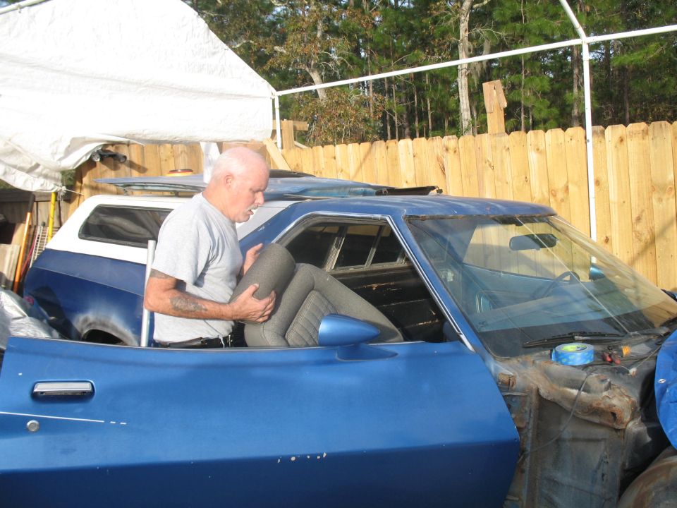 man holding car seat, blue car in foreground, blue and white SUV behind it